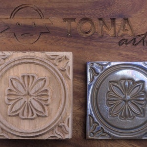 Könnte beinhalten: Zwei quadratische Fliesen mit einem geschnitzten Blumenmuster. Die Fliese links ist aus Holz und hat eine natürliche Oberfläche. Die Fliese rechts ist aus Keramik und hat eine dunkelblaue Glasur.