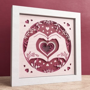 May include: White frame with a pink paper cutout design of two hands forming a heart shape around a layered heart. The design is surrounded by floral elements and small hearts.