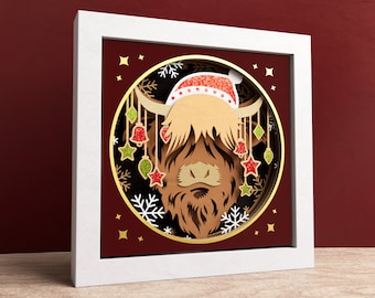May include: A white framed papercraft artwork featuring a Highland cow wearing a red Santa hat with white trim and gold bells. The cow is surrounded by snowflakes and stars.