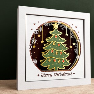 K&ouml;nnte beinhalten: Ein wei&szlig;er Rahmen mit einer goldenen und gr&uuml;nen Papierschnitt-Weihnachtsbaum-Darstellung in einem Kreis mit Schneeflocken und Sternen. Der Text "Merry Christmas" befindet sich am unteren Rand des Rahmens.