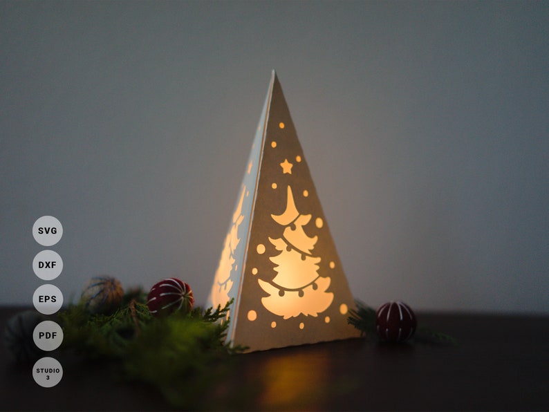 May include: A white paper Christmas tree lantern with a cut-out design of a Christmas tree with lights and ornaments. The lantern is lit from inside, casting a warm glow. The lantern is sitting on a wooden surface with green pine branches and red ornaments.