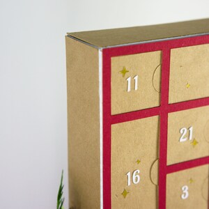 May include: Close-up of a cardboard advent calendar with a red frame. The calendar has individual doors numbered 3, 11, 12, 16, 19, and 21. Each door is a light brown color with a white number and gold star accents.