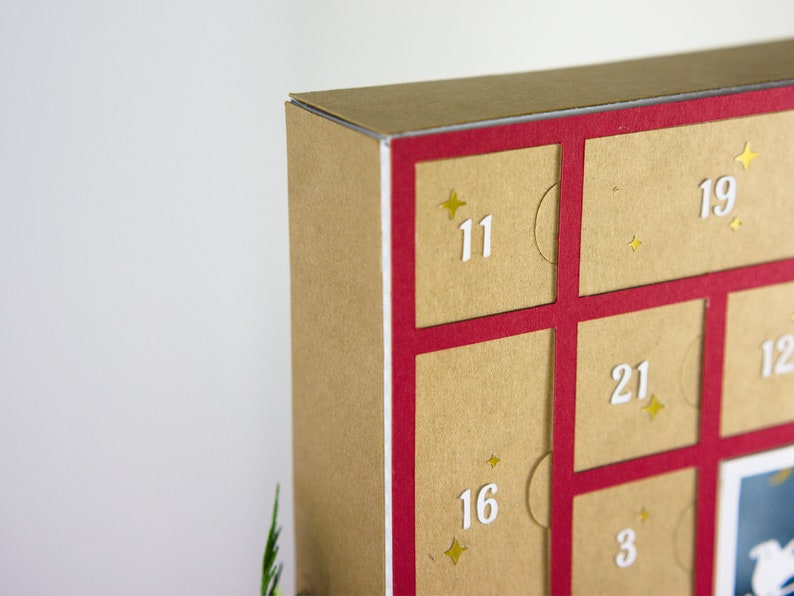 May include: Close-up of a cardboard advent calendar with a red frame. The calendar has individual doors numbered 3, 11, 12, 16, 19, and 21. Each door is a light brown color with a white number and gold star accents.