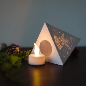 May include: A white paper lantern shaped like a triangle with a gold design of a moon, stars, and a snowflake. A small white tea light candle is lit inside the lantern.