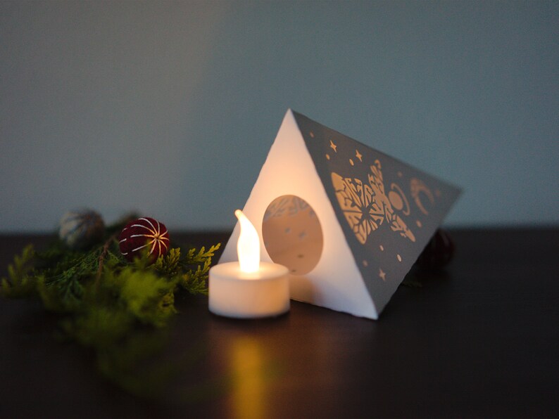 May include: A white paper lantern shaped like a triangle with a gold design of a moon, stars, and a snowflake. A small white tea light candle is lit inside the lantern.