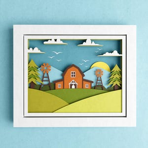 May include: A white frame with a paper cut-out design of a red barn with a white door and windows, two windmills, and green trees. The background is a blue sky with white clouds and a yellow sun.