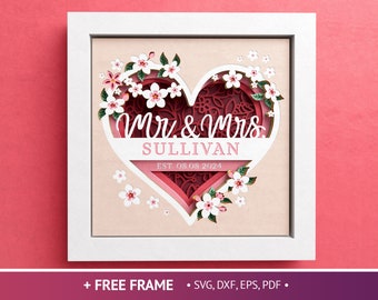 May include: A white frame with a pink background and a layered paper cut design of a heart with the text "Mr. & Mrs. SULLIVAN EST. 08.08.2024" inside. The heart is surrounded by pink flowers.