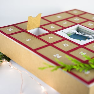 May include: A Christmas advent calendar with a kraft paper exterior and a red border. The calendar has numbered compartments, with one open to reveal a festive scene. Greenery and fairy lights add to the holiday theme.