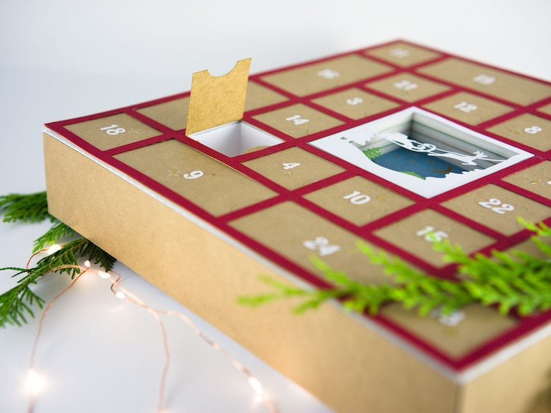 May include: A Christmas advent calendar with a kraft paper exterior and a red border. The calendar has numbered compartments, with one open to reveal a festive scene. Greenery and fairy lights add to the holiday theme.