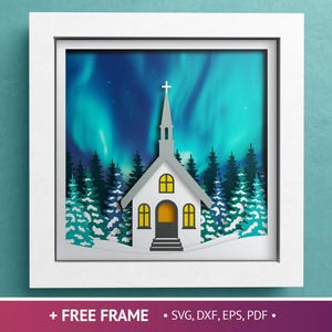 Könnte beinhalten: Gerahmtes Papierkunstwerk mit einer weißen Kirche mit Kreuz vor dem Hintergrund eines farbenprächtigen Polarlichts. Die Szene umfasst silhouettierte immergrüne Bäume und eine verschneite Landschaft. Der Rahmen ist weiß.