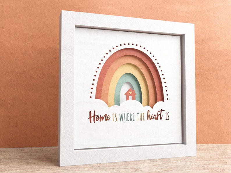 May include: White framed wall art with a rainbow design and the text "Home is where the heart is". The rainbow is made of layered paper in shades of red, orange, yellow, green, and blue. A small house is cut out of the center of the rainbow.