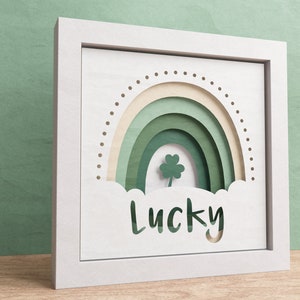 May include: White framed wall art with a green paper cut-out rainbow and a four-leaf clover. The word "Lucky" is cut out below the rainbow.