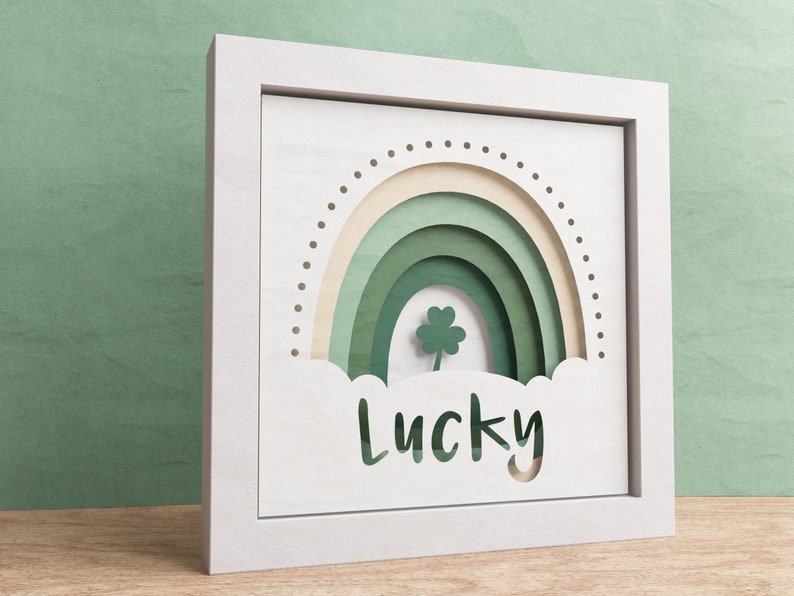 May include: White framed wall art with a green paper cut-out rainbow and a four-leaf clover. The word "Lucky" is cut out below the rainbow.