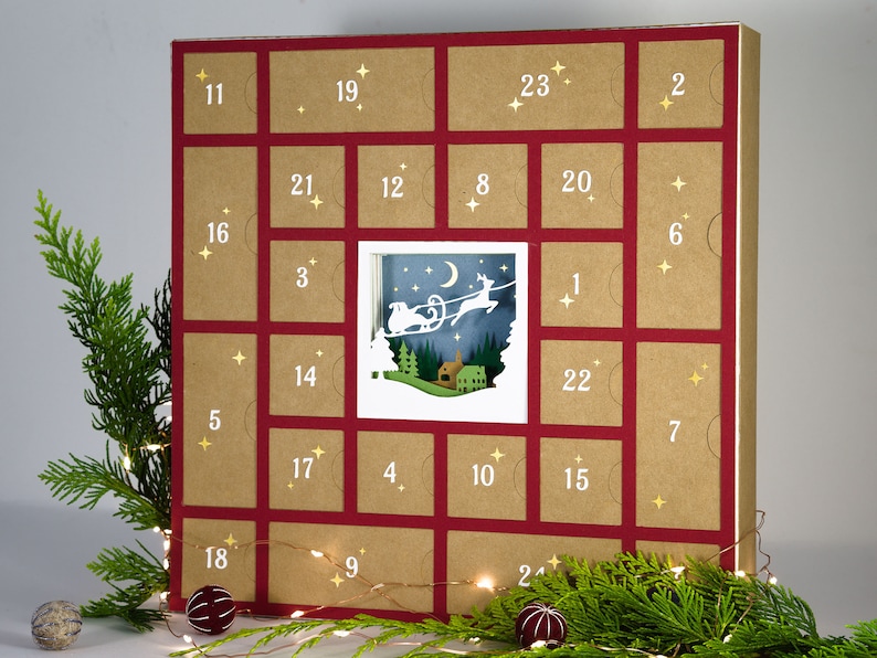 May include: A Christmas advent calendar with 24 numbered doors. The calendar is made of brown cardboard with red borders. A central window reveals a winter scene with a reindeer-pulled sleigh. Gold star accents and greenery decorate the calendar.