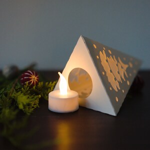 May include: A white paper Christmas tree lantern with a cut-out design of a tree and snowflakes. A small white LED tea light is placed in front of the lantern.