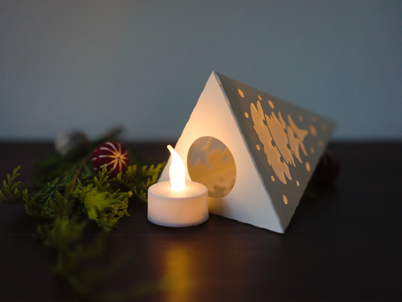 May include: A white paper Christmas tree lantern with a cut-out design of a tree and snowflakes. A small white LED tea light is placed in front of the lantern.