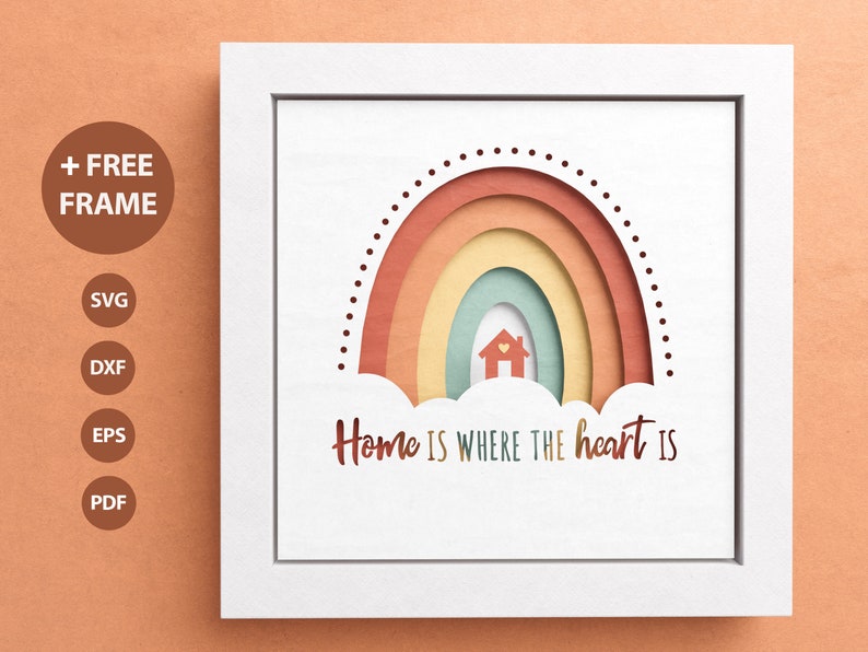 May include: A white frame with a white background and a rainbow with a house in the middle. The rainbow is made of paper and has a layered effect. The text "Home is where the heart is" is written below the rainbow.