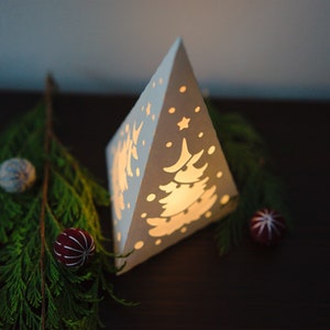 May include: A white paper pyramid lantern with a cut-out design of a Christmas tree and snowflakes. The lantern is lit from inside, casting a warm glow. The lantern is sitting on a dark brown surface with a sprig of evergreen branches and blue and red ornaments.