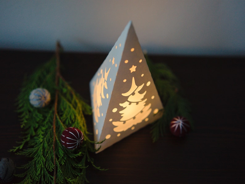 May include: A white paper pyramid lantern with a cut-out design of a Christmas tree and snowflakes. The lantern is lit from inside, casting a warm glow. The lantern is sitting on a dark brown surface with a sprig of evergreen branches and blue and red ornaments.