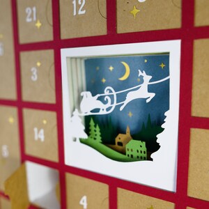 May include: A Christmas advent calendar with a red frame and individual brown envelopes. A white, layered paper cutout depicts Santa's sleigh and reindeer flying over a village under a starry night sky. Numbers 1-24 are printed on the envelopes.