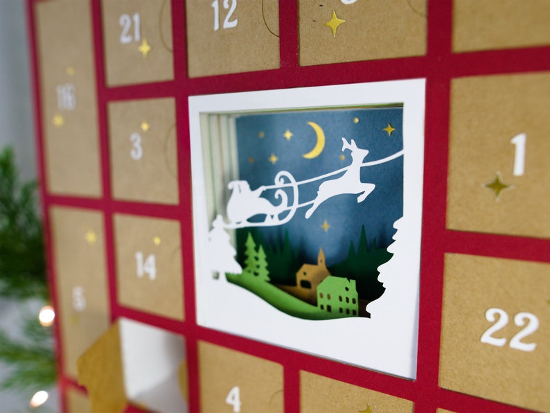 May include: A Christmas advent calendar with a red frame and individual brown envelopes. A white, layered paper cutout depicts Santa's sleigh and reindeer flying over a village under a starry night sky. Numbers 1-24 are printed on the envelopes.
