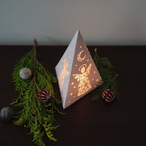 May include: White paper pyramid lantern with a cut-out design of an angel and a crescent moon. The lantern is lit from inside and casts a warm glow. The lantern is surrounded by small, round ornaments in shades of blue and red.
