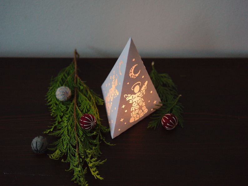 May include: White paper pyramid lantern with a cut-out design of an angel and a crescent moon. The lantern is lit from inside and casts a warm glow. The lantern is surrounded by small, round ornaments in shades of blue and red.