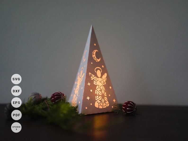 May include: A white paper Christmas tree with a cut-out design of an angel and a crescent moon. The tree is illuminated from within, casting a warm glow.