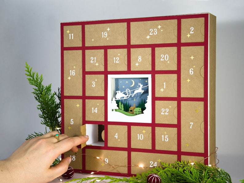 May include: A Christmas advent calendar with a red frame and individual doors numbered 1-24. The center door reveals a layered paper cut scene of a reindeer-pulled sleigh. A hand opens a door. Greenery and string lights are in the foreground.