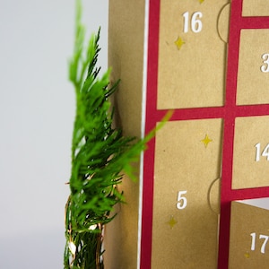 May include: A close-up of a Christmas advent calendar with a red border and tan pockets. The numbers 3, 5, 14, 16, and 17 are visible. Green foliage and small lights are on the left side of the image.