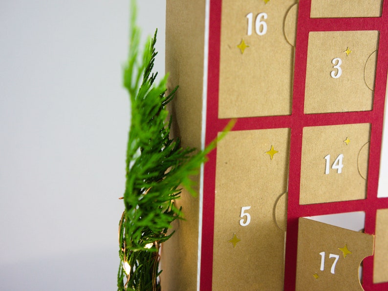 May include: A close-up of a Christmas advent calendar with a red border and tan pockets. The numbers 3, 5, 14, 16, and 17 are visible. Green foliage and small lights are on the left side of the image.