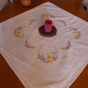 Könnte beinhalten: Weiße bestickte Tischdecke mit Blumenmuster und einer rosa Kerze auf einem Holzsockel in der Mitte.