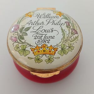 May include: A red and gold trinket box with a cream-coloured lid. The lid features the name "William Arthur Philip Louis" and the date "21st June 1982" surrounded by floral and crown designs. The box is round.