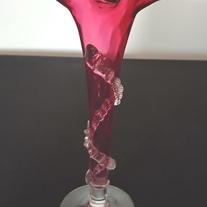 May include: A tall, ruby-red glass vase with a flared, ruffled top. A clear, decorative spiral adorns the vase's body. The vase has a circular base. The vase is likely used for decorative purposes.