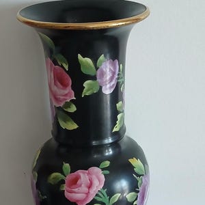 May include: A black vase with a flared rim featuring a gold-toned edge. The body of the vase is decorated with hand-painted pink and purple roses, accompanied by green leaves scattered across the surface. The vase has a rounded base and neck.