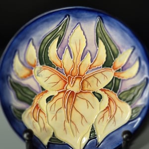 May include: A circular ceramic plate with a raised floral design. The plate has a blue background with a yellow and orange flower in the centre, surrounded by green leaves. The plate is displayed on a black stand.
