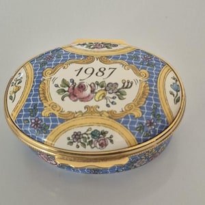 May include: An oval trinket box with a blue and white patterned lid. The lid features the year "1987" and floral accents in pink, yellow, and green. The box has a gold-coloured trim.