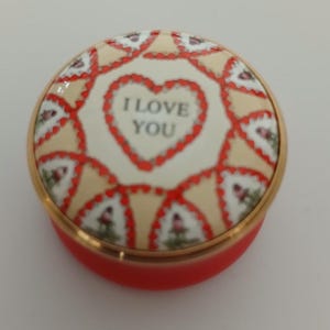 May include: A small, round trinket box in red with a gold rim. The lid displays a heart design with the text "I LOVE YOU". The lid also has a repeating pattern of red and white shapes, a charming gift.