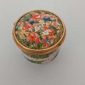 May include: A small, round trinket box with a floral design on the lid. The lid features a colourful arrangement of red, yellow, and blue flowers against a green background. The box has a gold-coloured rim and a green and pink striped body.