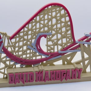 May include: A tan and magenta roller coaster model featuring a lattice support structure and a curved track. The base of the model displays the text DAVID MAHOGANY in raised magenta lettering against a tan background, with grey accents on the track.