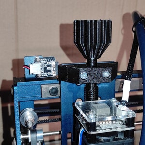 May include: Close-up of a blue and black 3D printer with various components. The printer features a black nozzle, a circuit board, and a small display with the text "2203". The design includes screws and a belt-driven system.