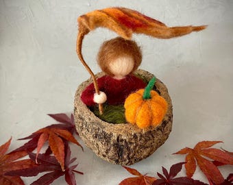 Op de afbeelding: Een gevilte herfstscène met een figuur in een rustieke kom, die een bladerenparaplu vasthoudt. Een oranje pompoen staat naast de figuur. Rode en oranje bladeren zijn rond de kom verspreid.
