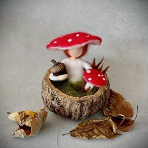 Könnte beinhalten: Eine Filz-Feenpuppe mit einem roten und weißen Pilz-Hut sitzt in einem ausgehöhlten Baumstamm mit einem kleinen roten und weißen Pilz und einer Eichel.