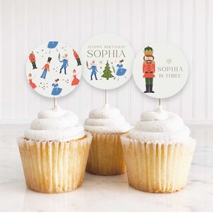 May include: Three white cupcakes with white frosting topped with round, colorful paper cupcake toppers featuring a Nutcracker theme. The toppers have illustrations of a Nutcracker, a Christmas tree, and dancing figures. The text on the toppers reads "Happy Birthday Sophia", "Sophia is Three", and a repeating pattern of Nutcracker figures.