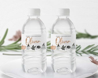 May include: Two clear plastic water bottles with personalized labels for a Halloween party. The labels feature a cute cartoon design with a spider, ghost, and cat. The text on the labels reads "Happy 2nd Birthday Oliver".