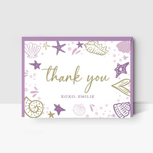 May include: A rectangular thank you card with a lavender border and a white background. The card features gold script text that says "thank you" and the name "Emilie". The card is decorated with seashell and starfish illustrations in gold and purple.