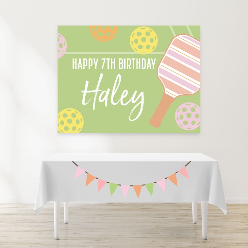 Op de afbeelding: Een groene en witte verjaardagsbord met de tekst "Happy 7th Birthday Haley" en een roze en oranje paddlebalracket. De banner is versierd met gele, oranje en roze paddleballen.
