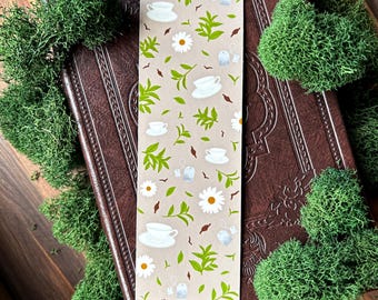 May include: A beige bookmark with a tea-themed design featuring white teacups, daisies, tea bags, and green leaves. The bookmark is rectangular and rests on a brown leather-bound book, surrounded by green moss.
