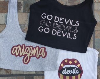 Può includere: Tre top corti con diversi design. Il primo è grigio con la parola "arizona" in lettere dorate e marroni. Il secondo è nero con le parole "Go Devils" ripetute tre volte in bianco. Il terzo è bianco con un design di labbra rosse e dorate con la parola "devils" all'interno.