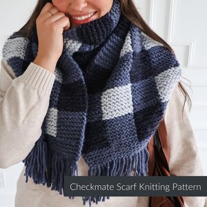 May include: A chunky knit scarf featuring a checkered pattern in shades of navy blue, slate blue, and light gray. The scarf has a thick ribbed collar and long fringe along the bottom edge. The text Checkmate Scarf Knitting Pattern is visible.